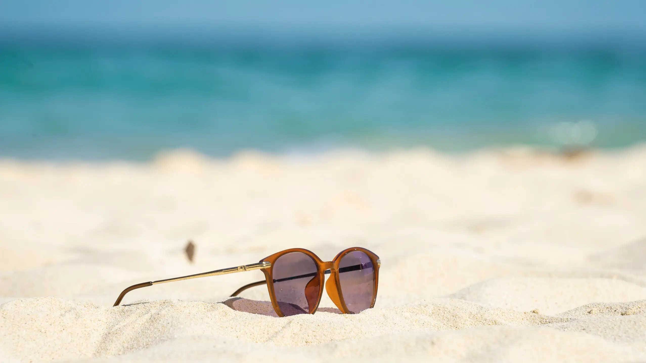 orangene Sonnenbrille liegt auf fast weissem Sand am Strand im Hintergrund türkises Meer