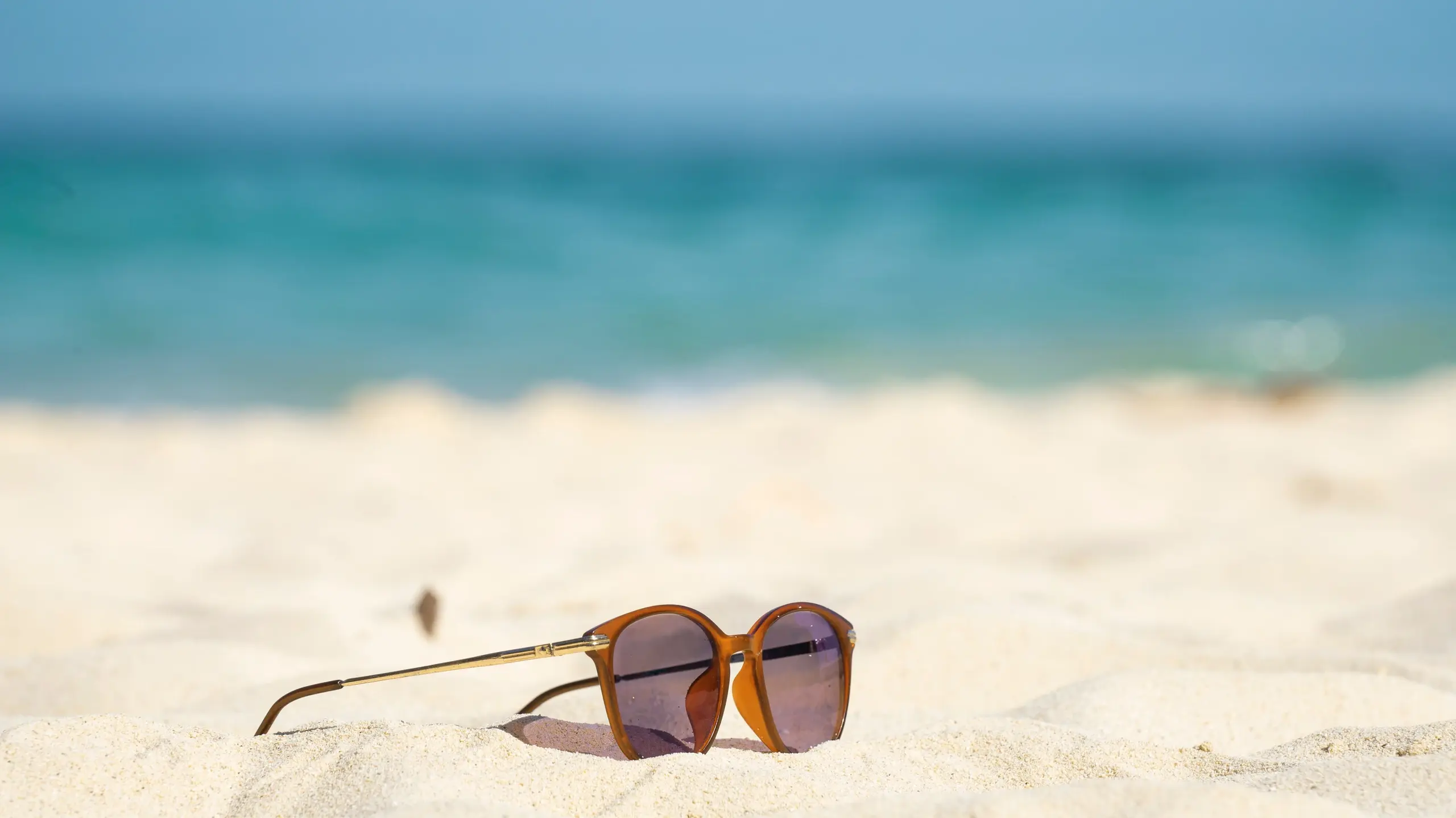 orangene Sonnenbrille liegt auf fast weissem Sand am Strand im Hintergrund türkises Meer