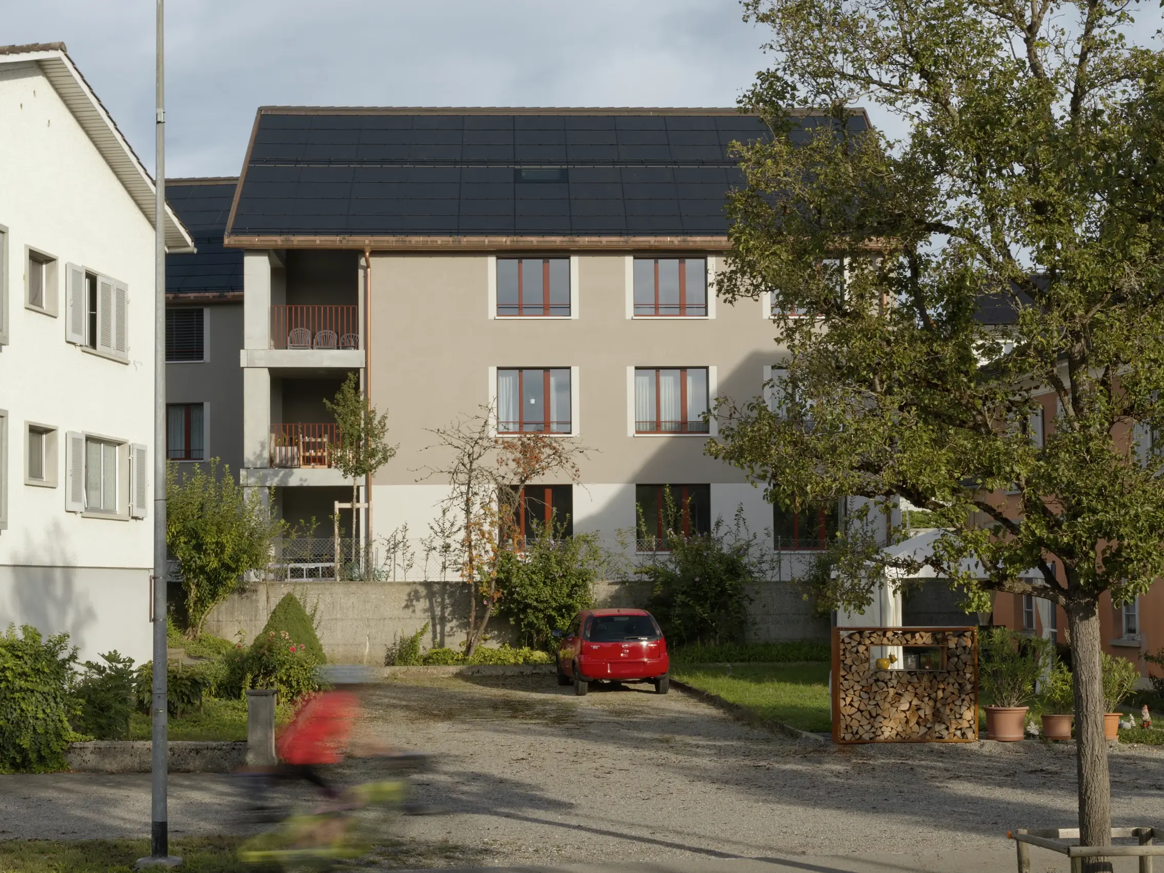 Vue du bâtiment depuis la rue ; une voiture rouge est garée devant et un arbre se trouve au premier plan