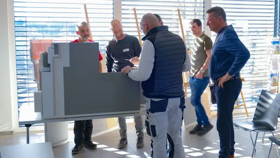 Mehrere Personen stehen um das grosse Muster für Fixit RoofEtics® und begutachten es genau, im Hintergrund Raff Stores und Sonnenlichtstreifen
