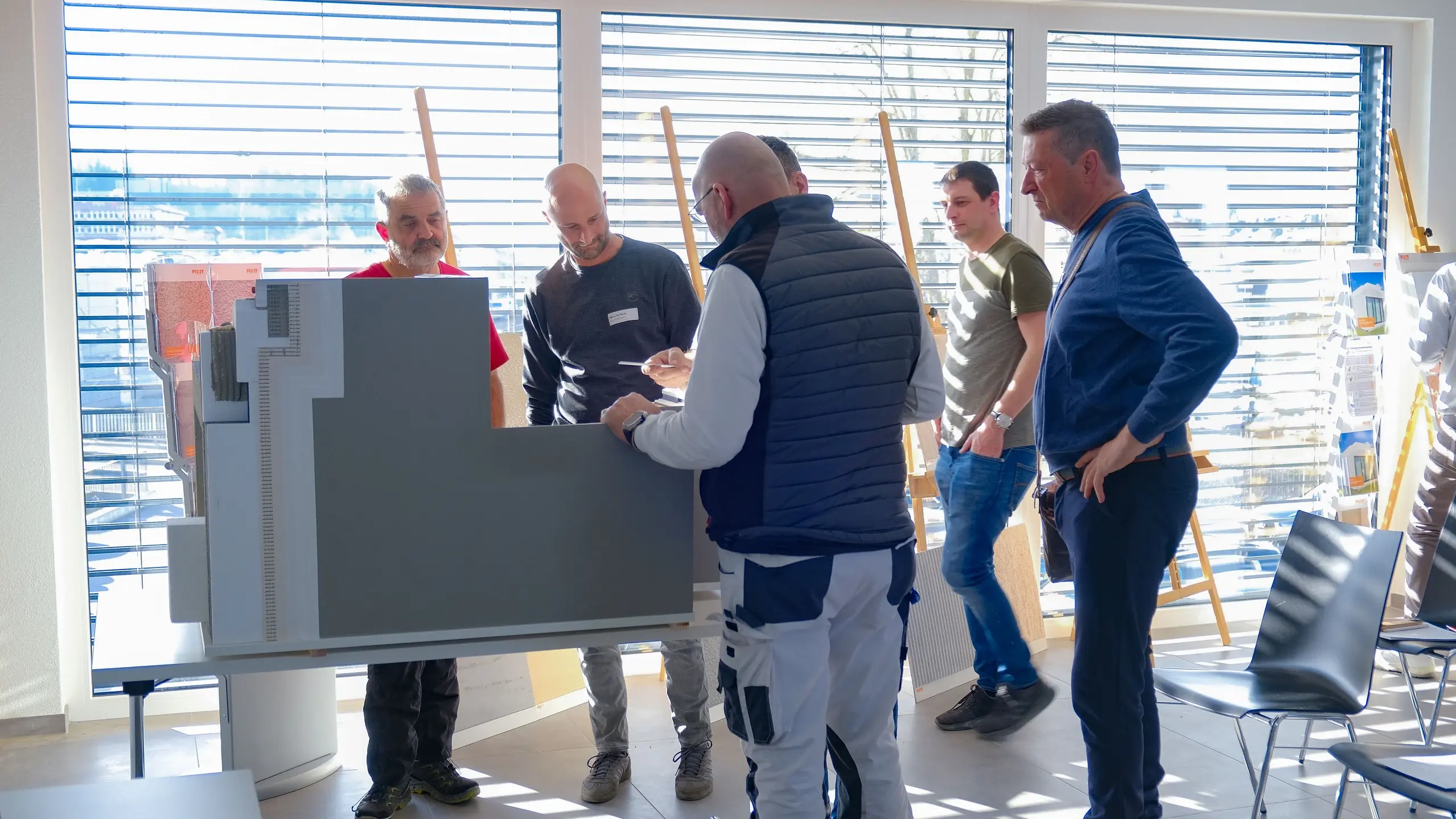 Mehrere Personen stehen um das grosse Muster für Fixit RoofEtics® und begutachten es genau, im Hintergrund Raff Stores und Sonnenlichtstreifen