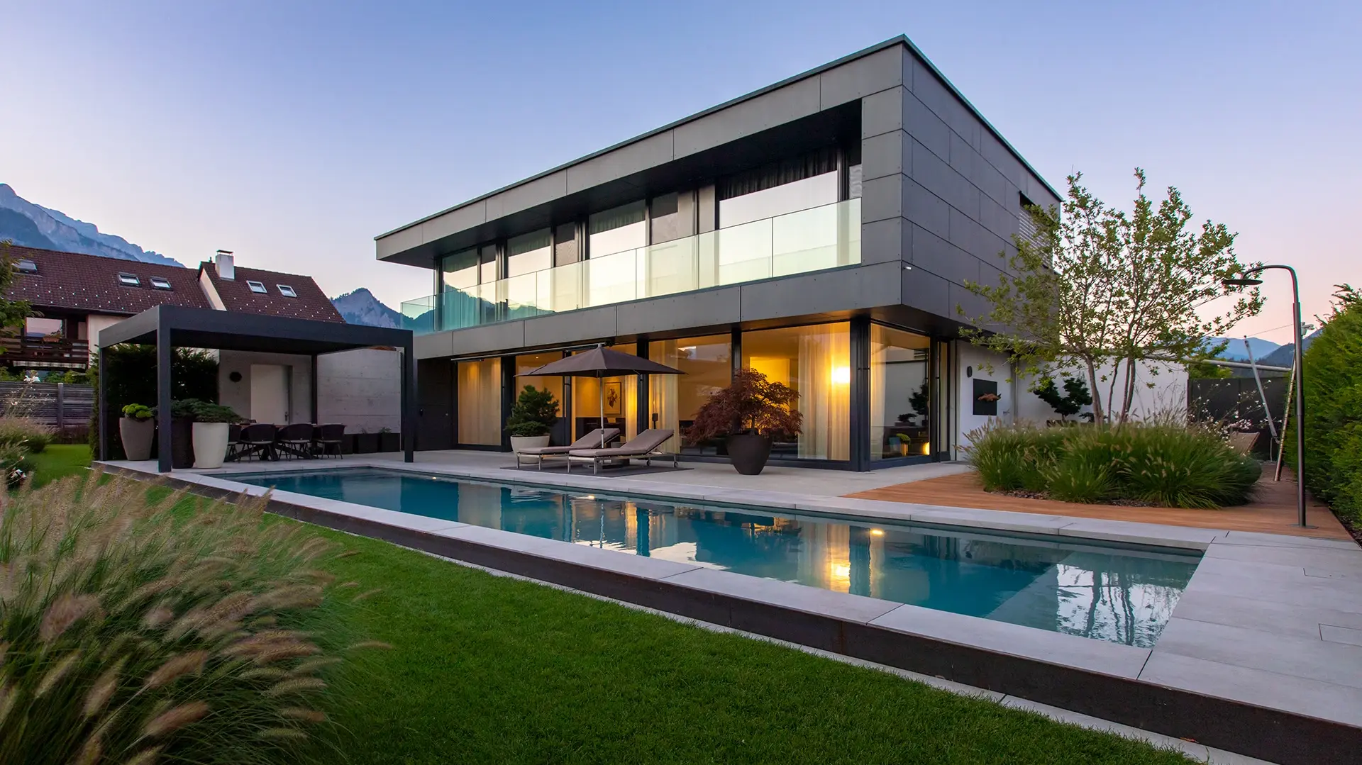 Une piscine rectangulaire baignée par la lumière du soir et une villa moderne à deux étages, dotée d'une façade en verre et en dalles
