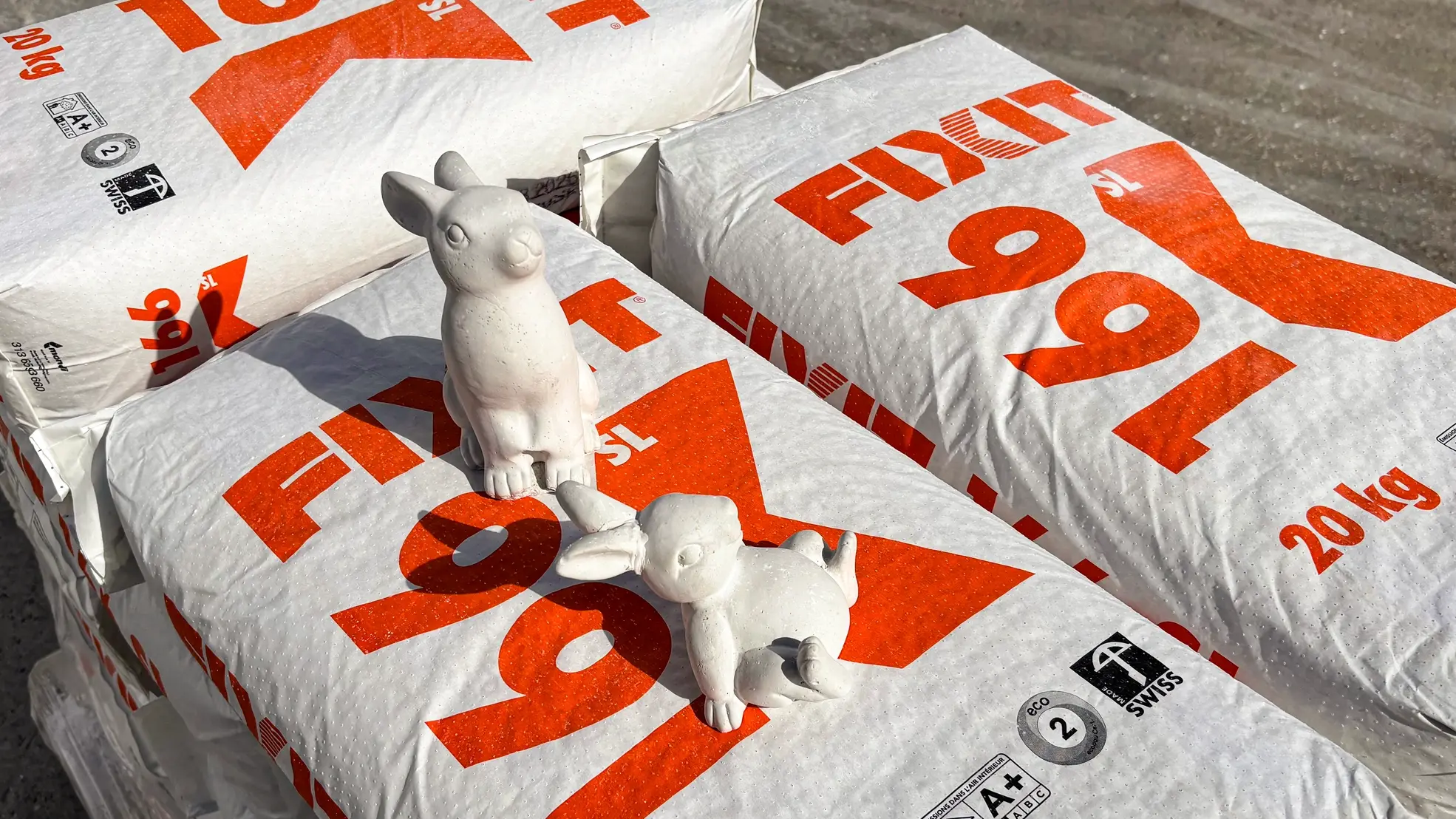 Säcke auf Palette mit Fixit 166 SL plus, zwei weisse Gips-Osterhasen sitzen darauf im Sonnenschein