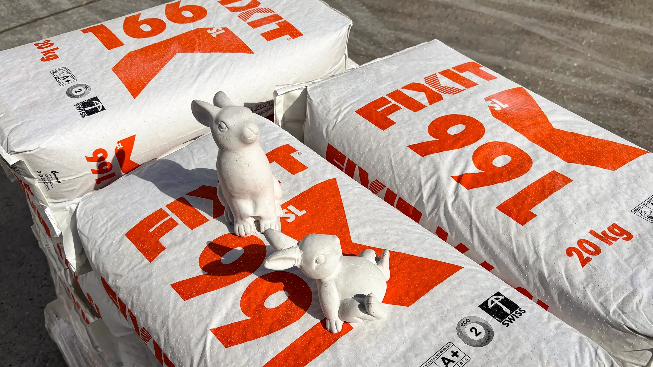 Säcke auf Palette mit Fixit 166 SL plus, zwei weisse Gips-Osterhasen sitzen darauf im Sonnenschein