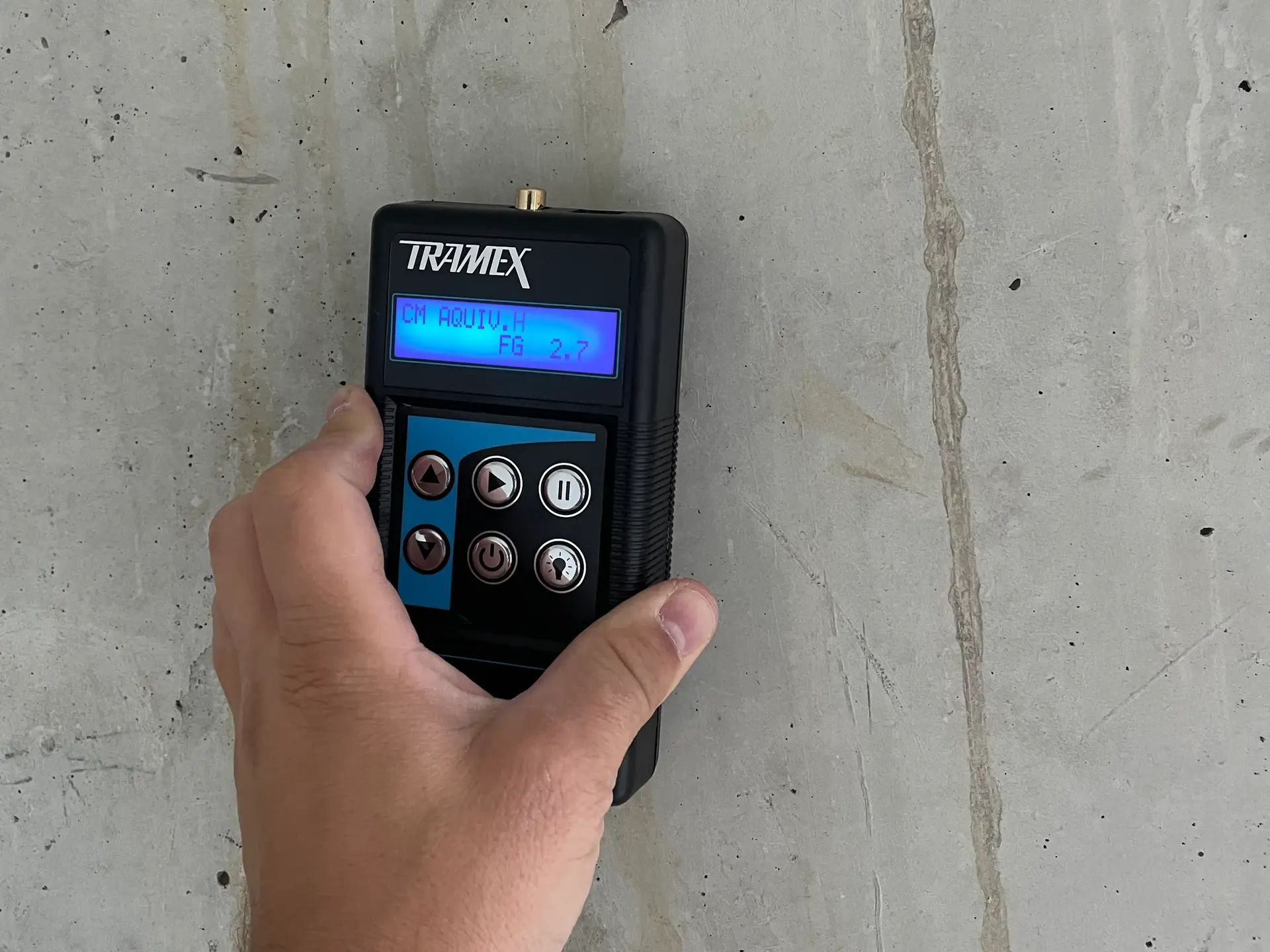 Eine Hand hält das Tramex Messgerät an eine trockene Betonwand, der angezeigte Wert beträgt 2.7