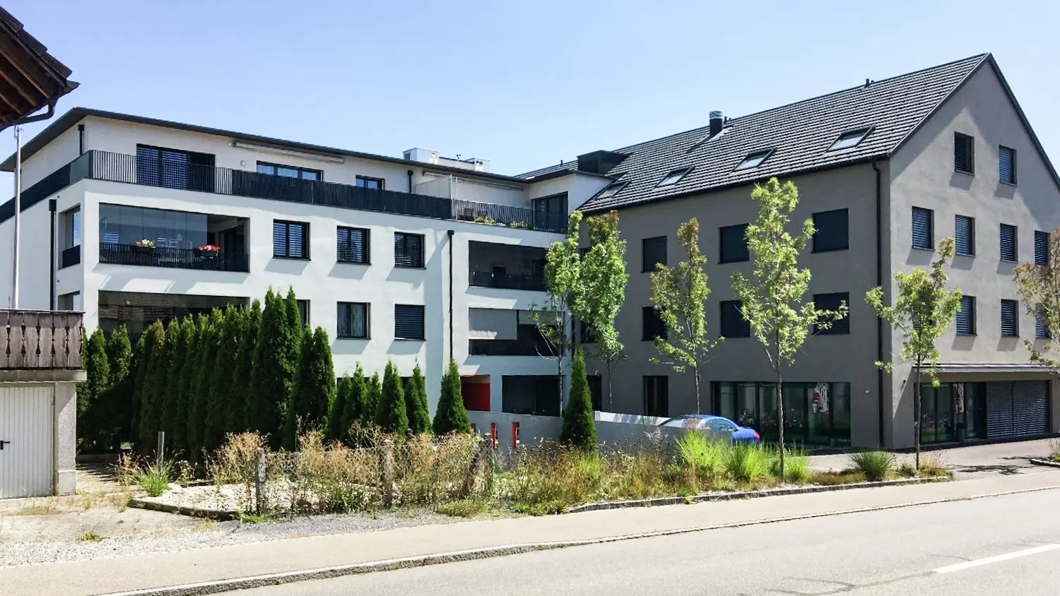 L-förmiges Gebäude flachem Dach und Giebeldach mit einigen Bäumen und Hecke im Vordergrund