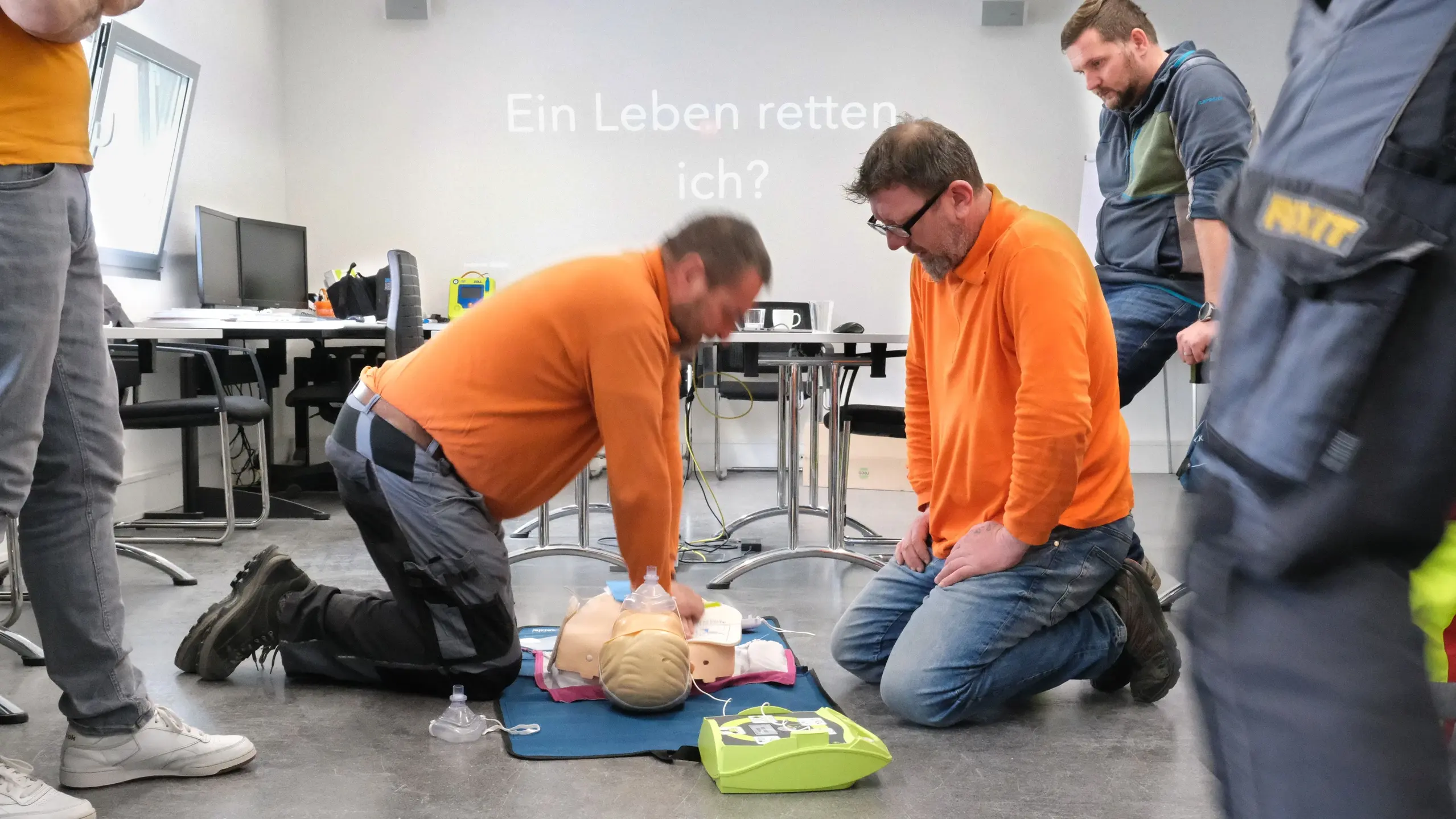 Zwei Fixit Mitarbeiter in orangenen Shirts üben die Reanimation an einer Puppe mit dem Defibrillator