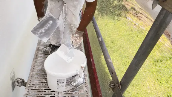 Arbeiter auf einem Gerüst mit einer weiss verputzten Fassade, er arbeitet mit zwei Kellen den neuen Fixit Silikatputz aussen aus dem Eimer auf die Fassade