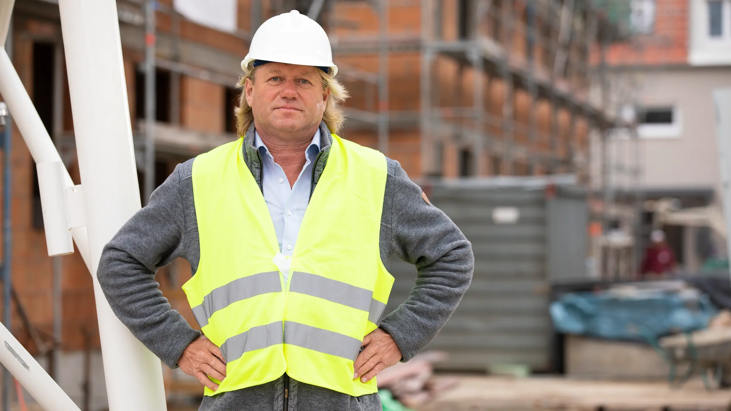 Hellhäutiger Mann mit mittellangen, blonden Haaren und einem weissen Bauhelm und gelber Warnweste steht mit den Händen in die Seiten gestützt vor einer Baustelle