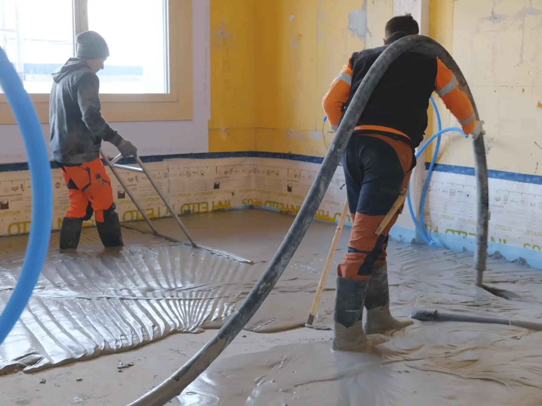 Ein Verarbeiter bringt den Fixit POR Schaummörtel mit dem Estrich-Schlauch über der Schulter als Ausgleichsschicht auf den Fussboden im Innenraum ein. Eine zweite Person schwabbelt den Bereich.