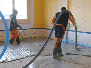 Un applicateur applique le mortier mousse Fixit POR avec le tuyau de chape au-dessus de l'épaule comme couche d'égalisation sur le sol à l'intérieur. Une deuxième personne balaie la zone.