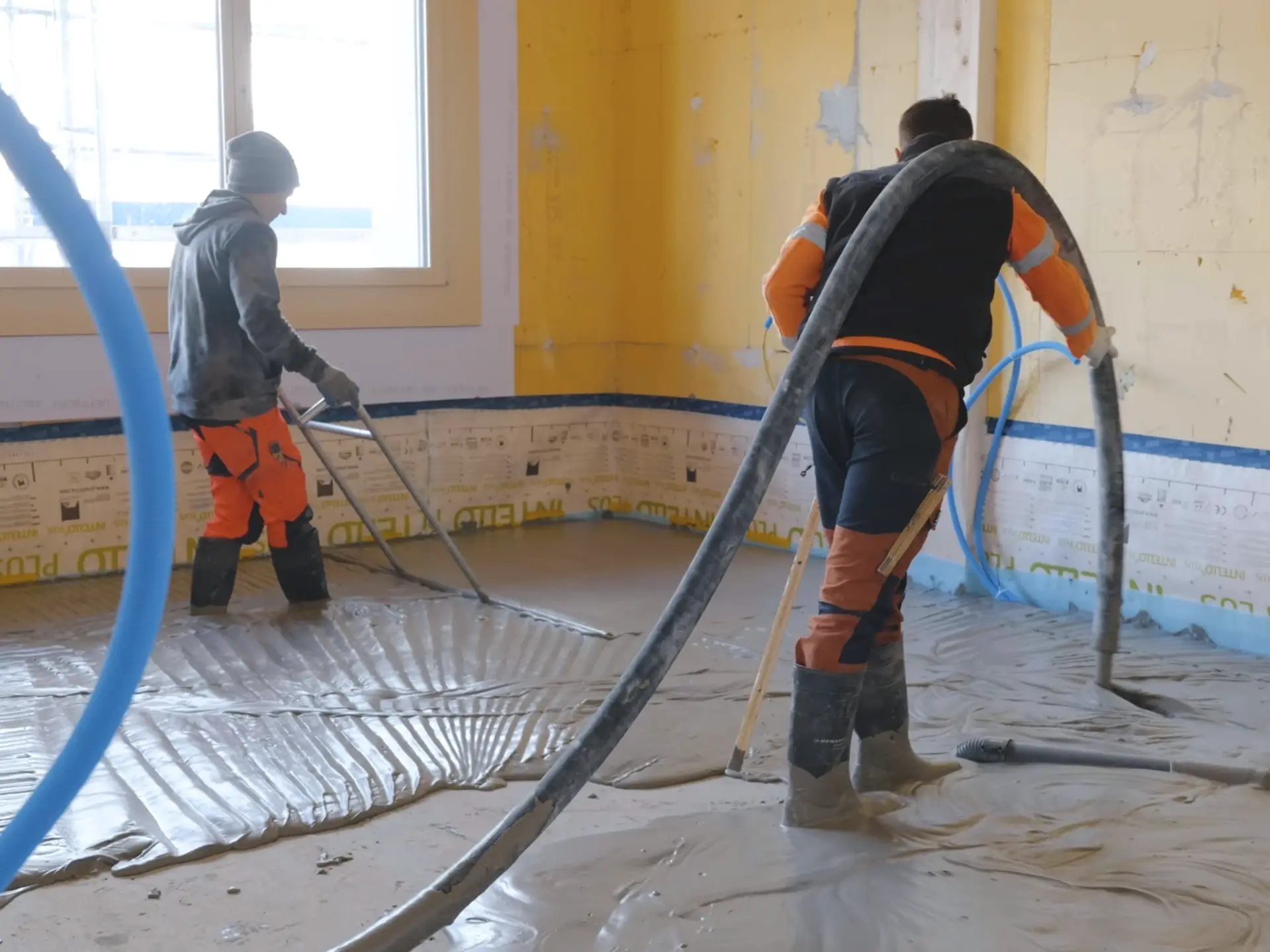 Ein Verarbeiter bringt den Fixit POR Schaummörtel mit dem Estrich-Schlauch über der Schulter als Ausgleichsschicht auf den Fussboden im Innenraum ein. Eine zweite Person schwabbelt den Bereich.