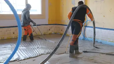 Ein Verarbeiter bringt den Fixit POR Schaummörtel mit dem Estrich-Schlauch über der Schulter als Ausgleichsschicht auf den Fussboden im Innenraum ein. Eine zweite Person schwabbelt den Bereich.