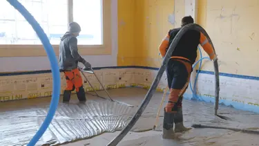 Un applicateur applique le mortier mousse Fixit POR avec le tuyau de chape au-dessus de l'épaule comme couche d'égalisation sur le sol à l'intérieur. Une deuxième personne balaie la zone.