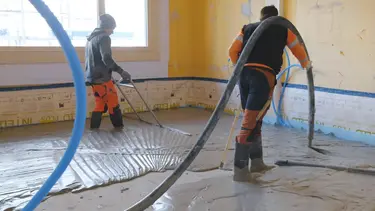 Un applicateur applique le mortier mousse Fixit POR avec le tuyau de chape au-dessus de l'épaule comme couche d'égalisation sur le sol à l'intérieur. Une deuxième personne balaie la zone.