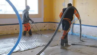 Ein Verarbeiter bringt den Fixit POR Schaummörtel mit dem Estrich-Schlauch über der Schulter als Ausgleichsschicht auf den Fussboden im Innenraum ein. Eine zweite Person schwabbelt den Bereich.