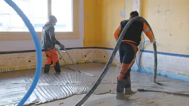 Un applicateur applique le mortier mousse Fixit POR avec le tuyau de chape au-dessus de l'épaule comme couche d'égalisation sur le sol à l'intérieur. Une deuxième personne balaie la zone.