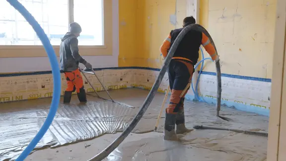 Un applicateur applique le mortier mousse Fixit POR avec le tuyau de chape au-dessus de l'épaule comme couche d'égalisation sur le sol à l'intérieur. Une deuxième personne balaie la zone.