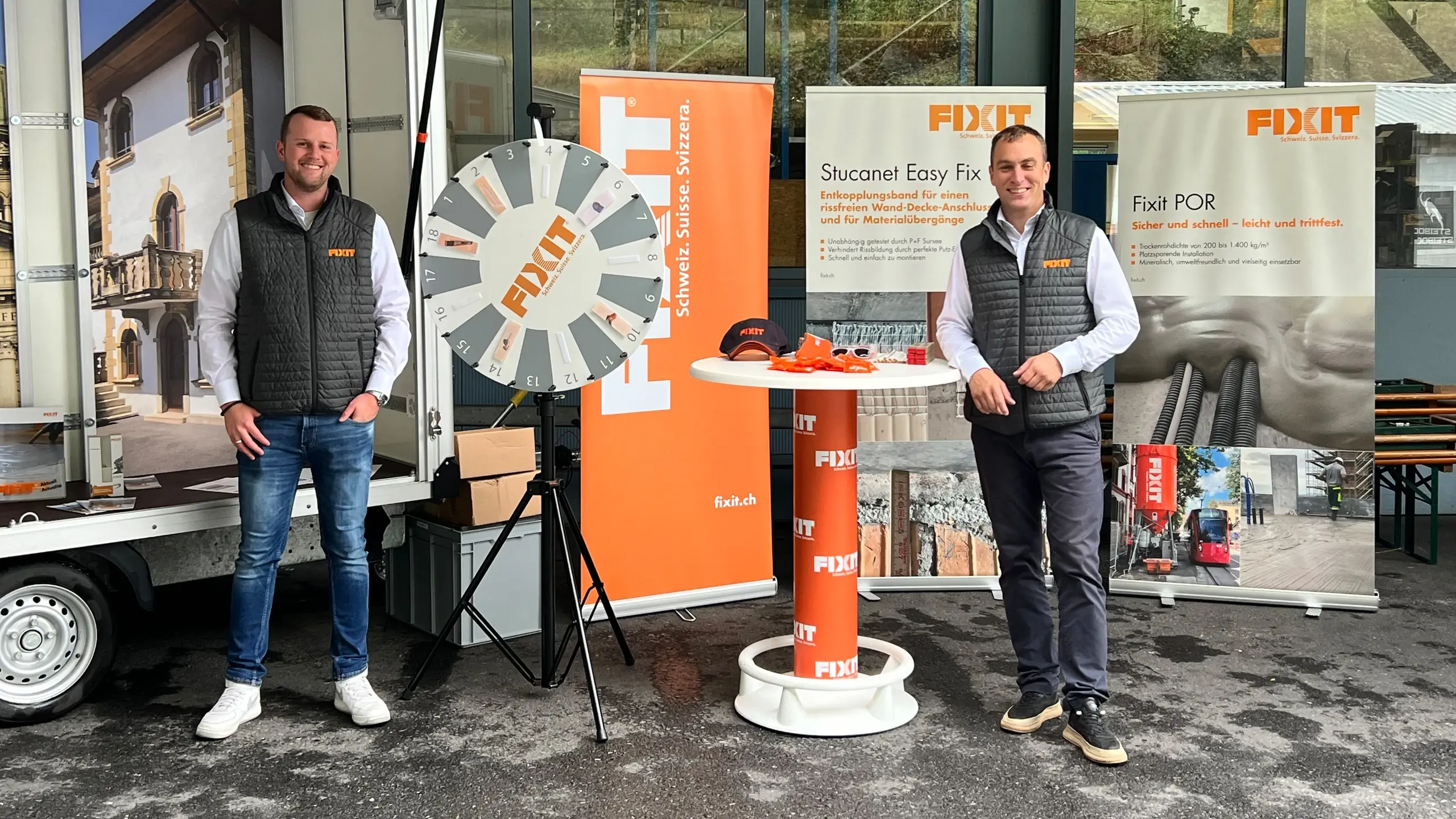 Fixit Mitarbeiter Thierry Lips (links) und Miroslav Rousar (rechts) vor dem Roadshow-Anhänger mit Fixit Glücksrad, Stehtisch und drei Werbebannern