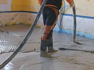 Un applicateur applique le mortier mousse Fixit POR avec le tuyau de chape au-dessus de l'épaule comme couche d'égalisation sur le sol à l'intérieur. Une deuxième personne balaie la zone.