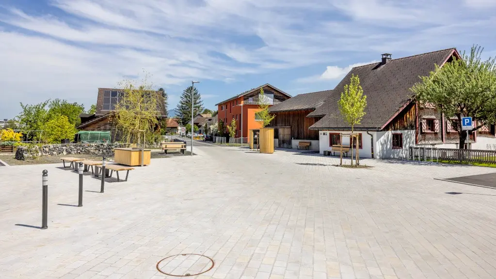 Pflästerung mit hellgrauen Steinen auf einem weitläufigen Dorfplatz, mehrere Einfamilienhäuser im Hintergrund