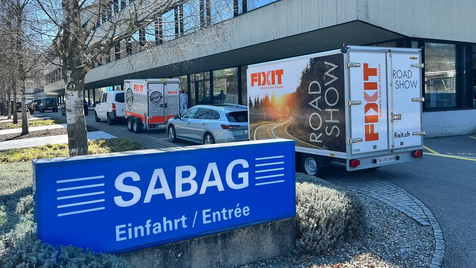 Blaues Schild mit SABAG Logo an der Einfahrt und im Hintergrund zwei Fahrzeuge mit jeweils einem Fixit Roadshowanhänger vor dem Bürogebäude