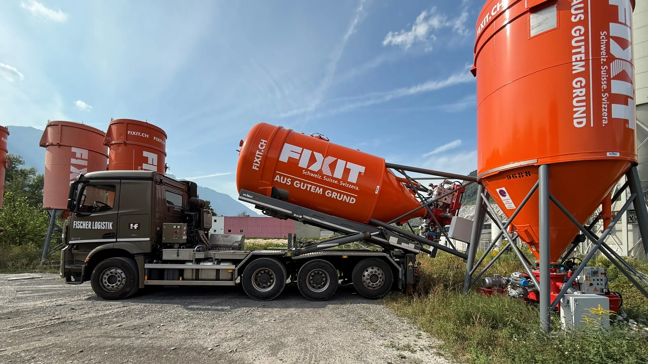 Brauner Fischer Logistik LKW läd ein orangenes Fixit Drucksilo mit CLEVER-Einheit auf einen Wiesenstellplatz ab