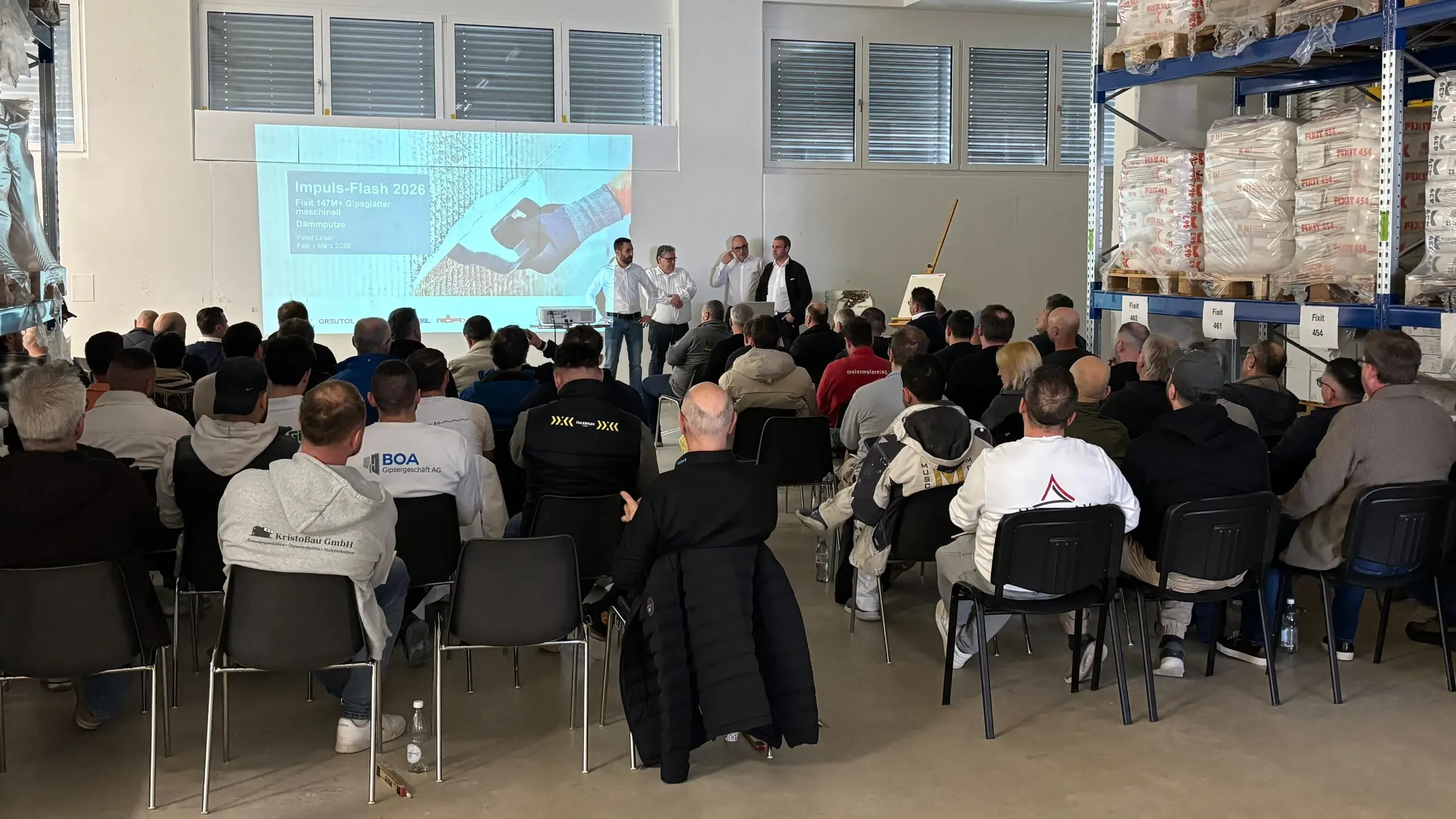 Raumansicht mit den Teilnehmenden und dem Intro-Slide der Präsentation sowie Ralph Herrman, Werner Wälle, Peter Loser und Marco Kern, die die Teilnehmenden begrüssen