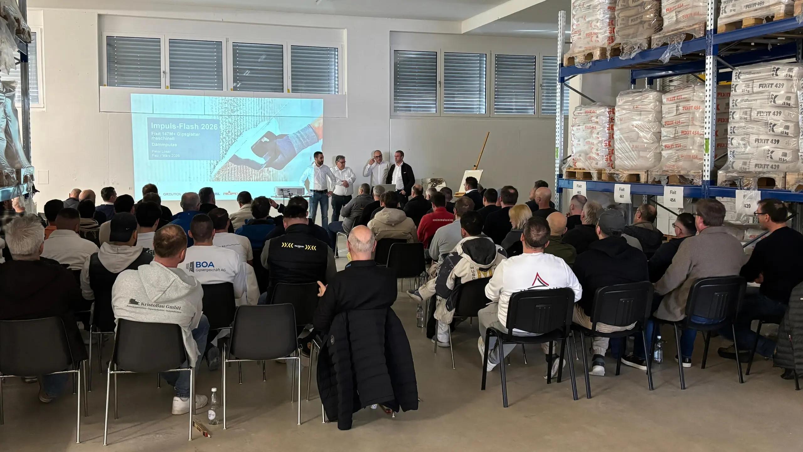 Raumansicht mit den Teilnehmenden und dem Intro-Slide der Präsentation sowie Ralph Herrman, Werner Wälle, Peter Loser und Marco Kern, die die Teilnehmenden begrüssen