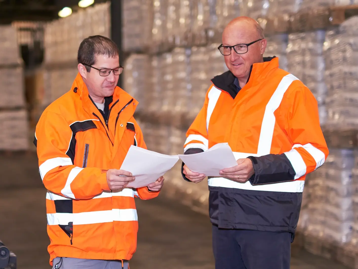 Two Fixit employees in the logistics department wearing high-visibility clothing stand in front of the product warehouse with Fixit bags and check delivery documents.