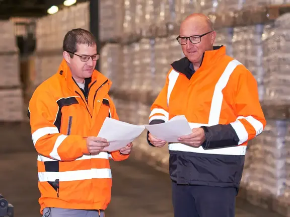Deux collaborateurs Fixit du secteur logistique en tenue de sécurité se tiennent devant l'entrepôt de produits avec des sacs Fixit et vérifient les documents de livraison.