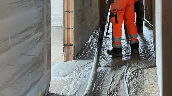 Vue détaillée d'un couloir en cours de construction avec un tuyau de chape, les jambes d'un ouvrier et du mortier mousse Fixit POR liquide sur le sol.