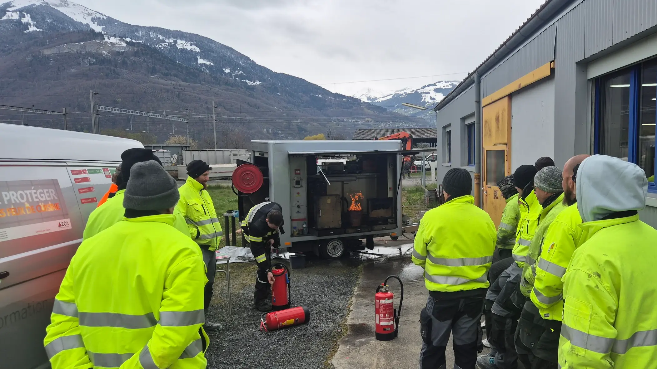 Mehrere Personen mit gelber Schutzkleidung stehen als Gruppe versammelt um den Schulungsanhänger vor den Feuerlöschern