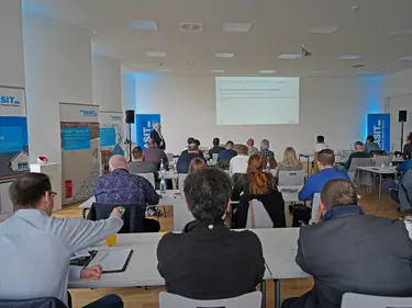  Eine Person hält eine Präsentation, die anderen hören aufmerksam zu, in einem Veranstaltungssaal.