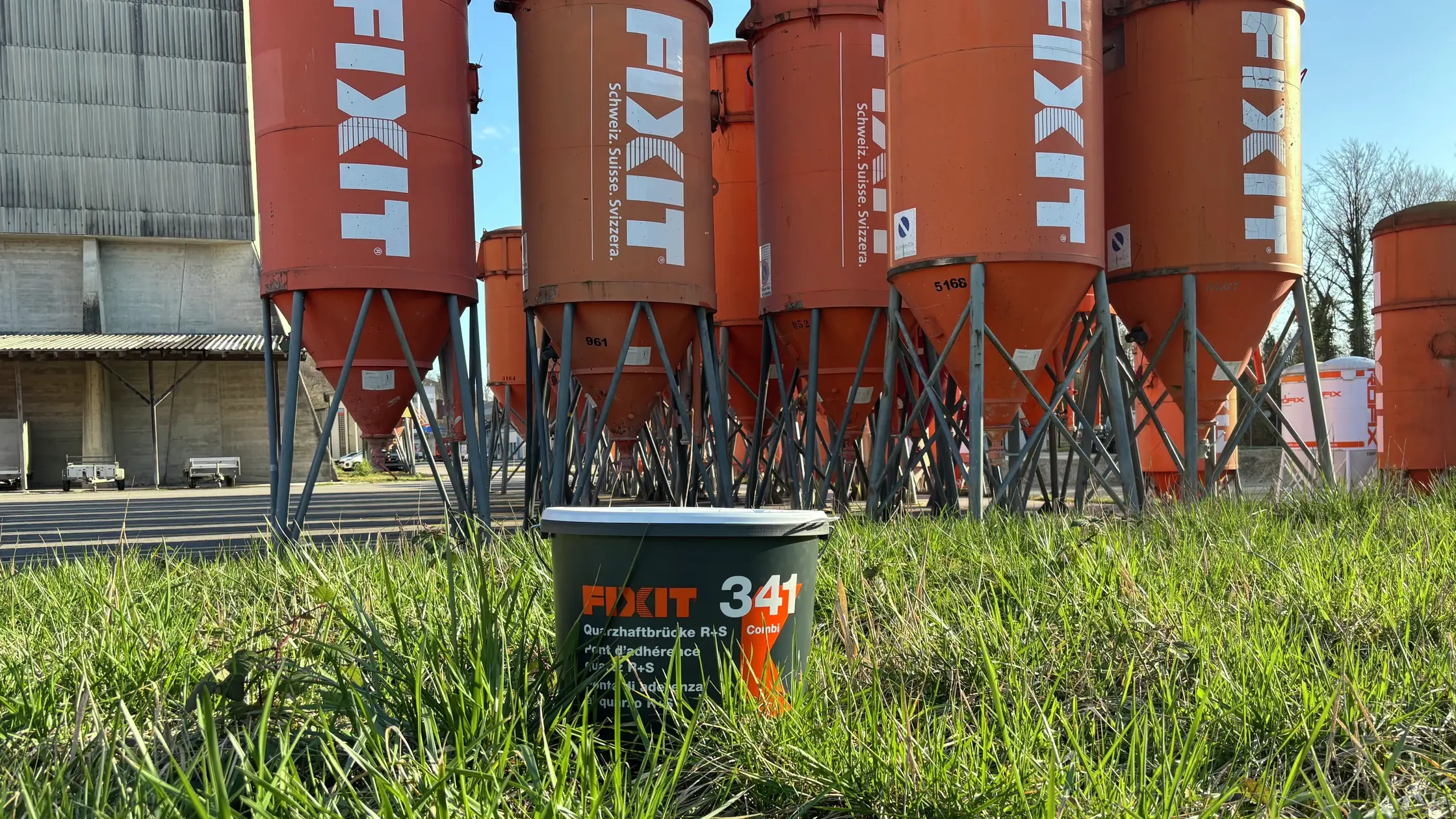 Fixit 341 im grauen Recycling-Eimer steht auf einer grünen Wiese im Sonnenschein vor orangenen Fixit Silos