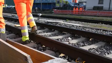 Zwei Bauarbeiter in orangener Arbeitskleidung laufen am Gleisbett mit dem Sichtschacht für die Verfüllung mit Fixit POR vorbei