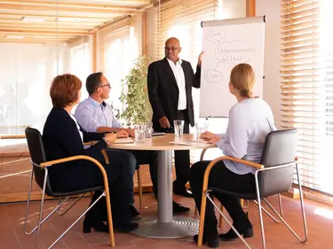 Eine Person präsentiert, und drei andere Personen am Tisch hören aufmerksam zu