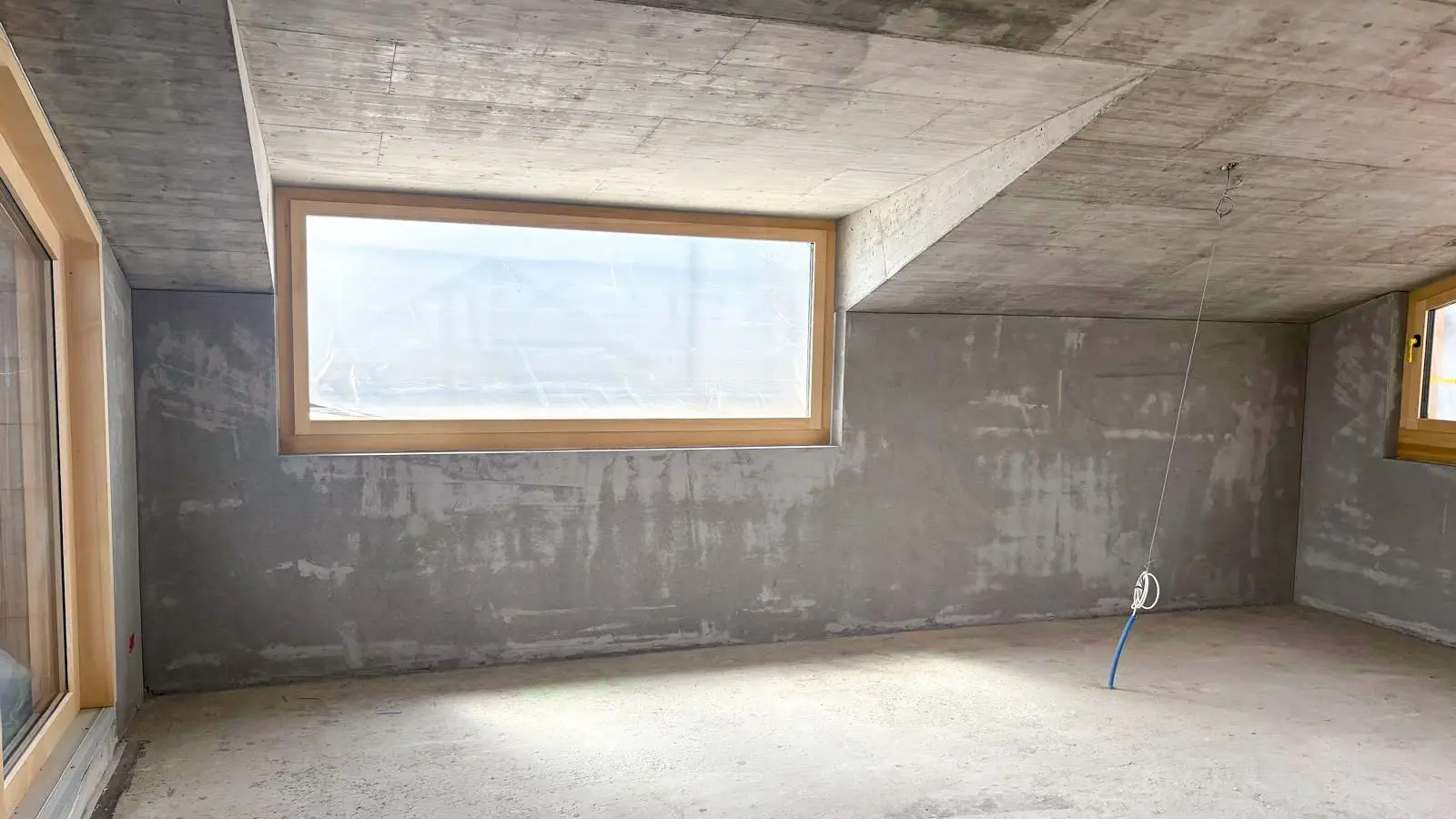 Raumansicht des Rohbaus mit Dachschräge und verputzten Wandflächen sowie einem grossen, länglichen, rechteckigen Fenster