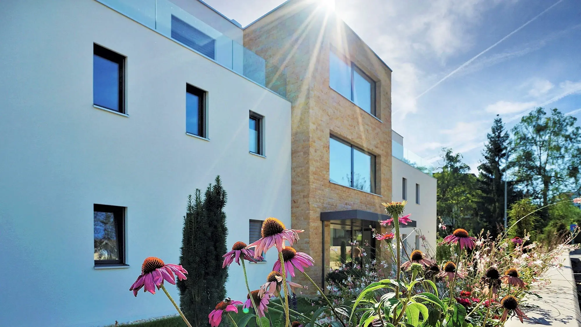 Mehrfamilienhaus mit einer Fassade aus Putz und Naturstein mit Blumen im Vordergrund und Sonnenstrahlen am oberen Bildrand