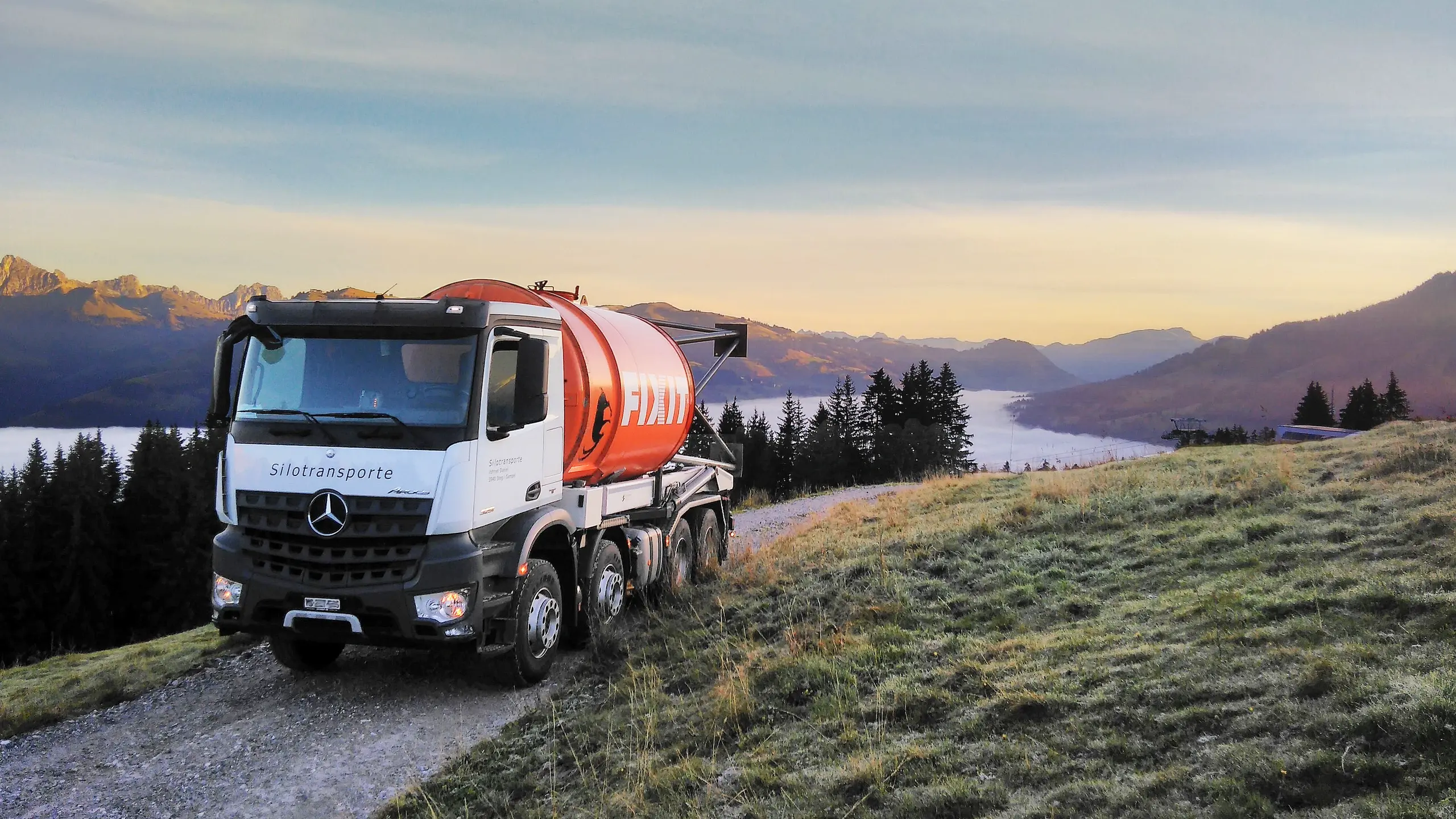 Camion bianco con un silo Fixit arancione su una strada sterrata in un altopiano, con le montagne sullo sfondo e la nebbia nella valle