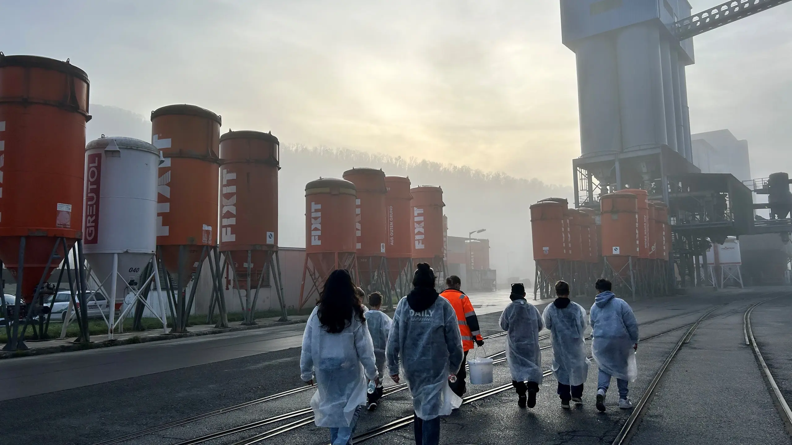 Kindergruppe von hinten in Schutzkleidung zwischen Silos im Nebel mit Sonnenaufgang auf dem Fixit Werksgelände
