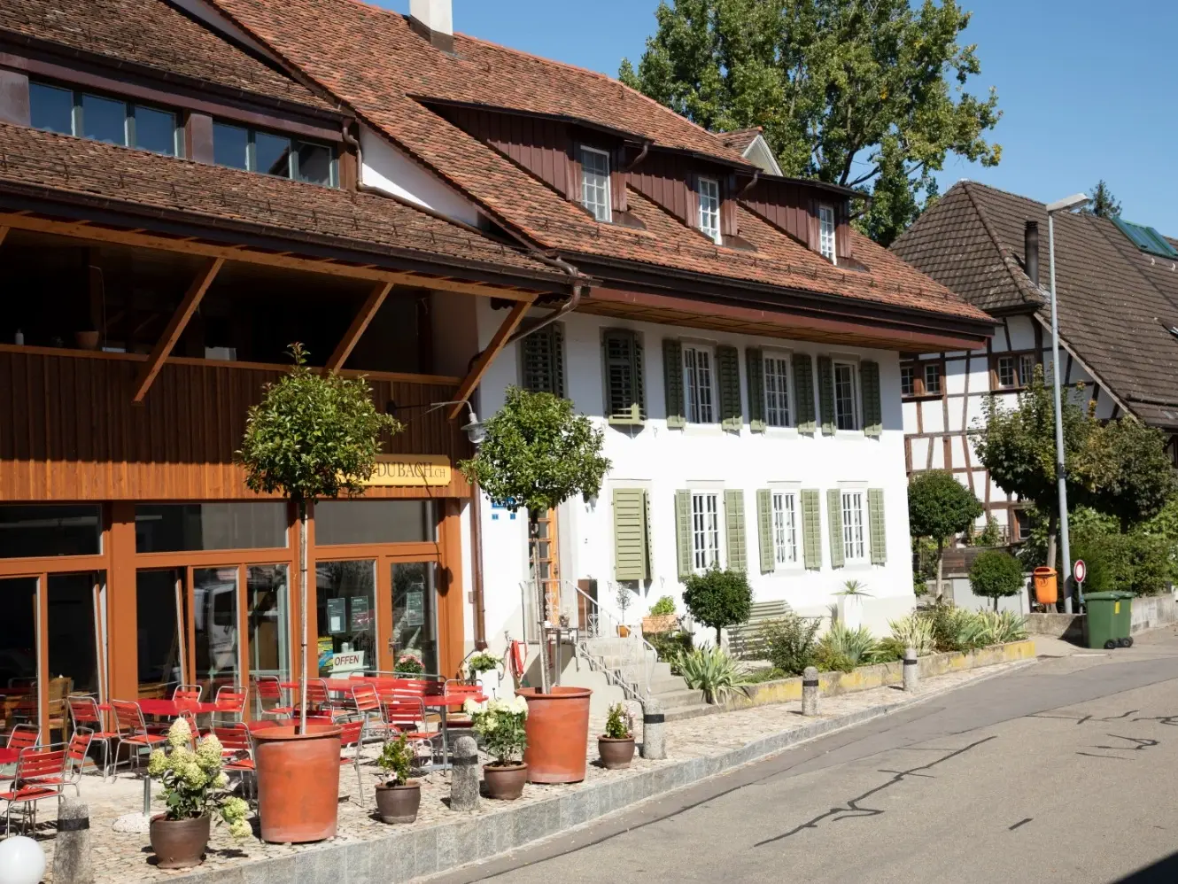 Zwei verbundene Gebäude, links ein dunkles mit Kaffeetischen und Holz und daran anschliessend ein weiss verputztes Gebäude mit grünen Fensterläden