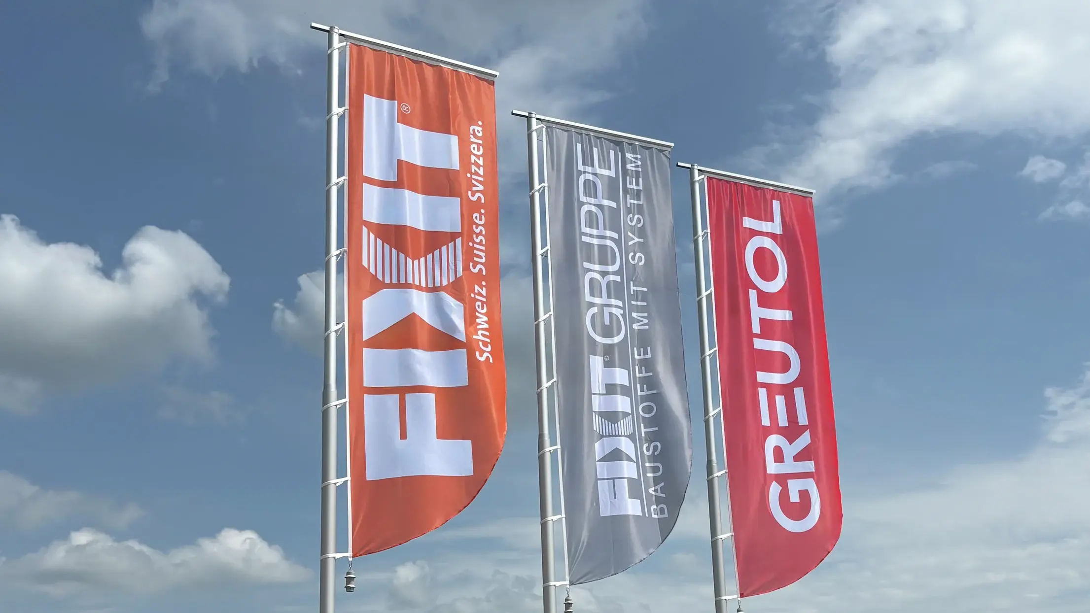Drei Fahnen vor blauem Himmel mit weissen Wolken, von links nach rechts orangene Fahne mit Fixit Logo, graue Fahne mit FIXIT GRUPPE Logo und rote Fahne mit Greutol Logo