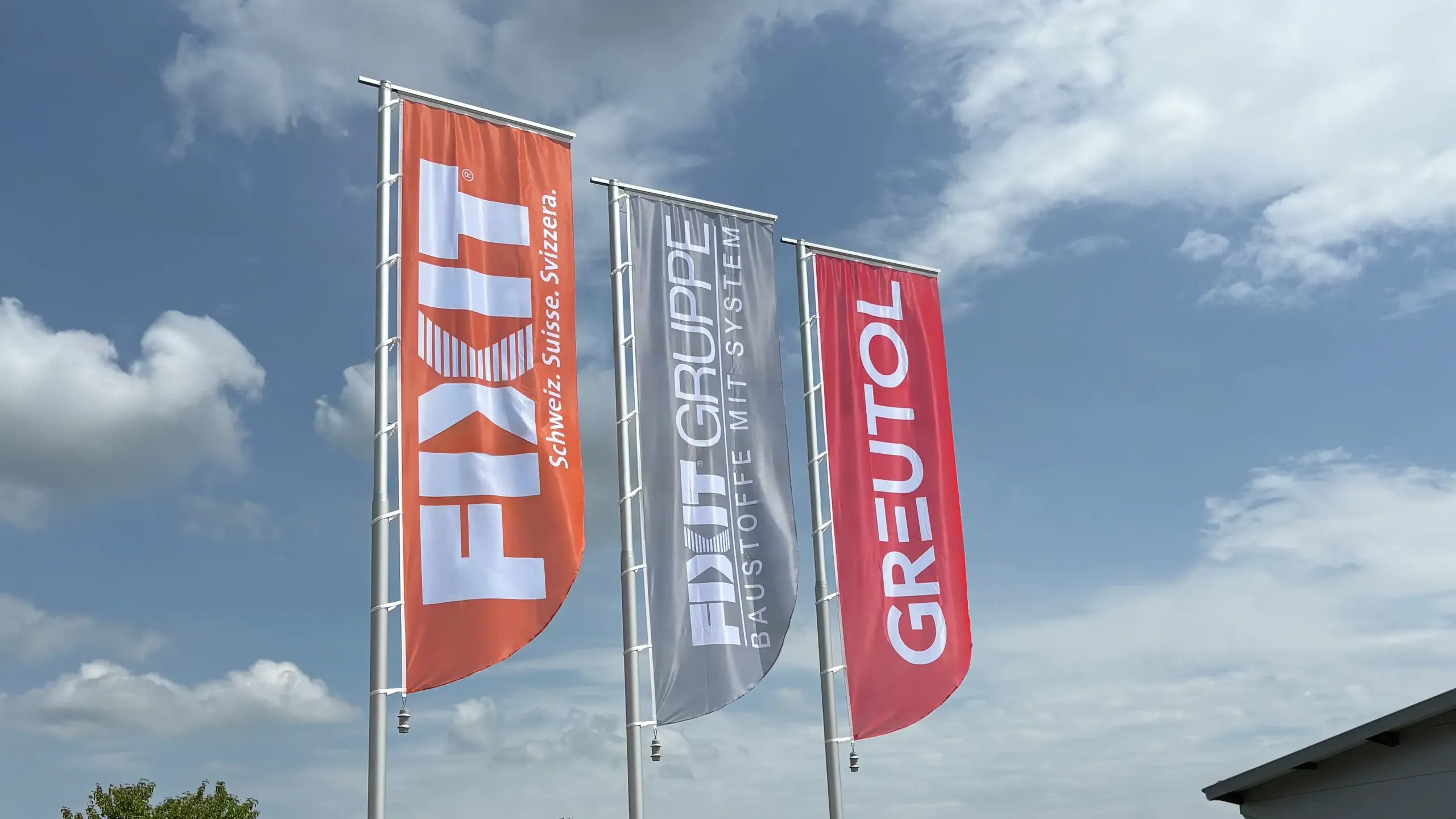 Drei Fahnen vor blauem Himmel mit weissen Wolken, von links nach rechts orangene Fahne mit Fixit Logo, graue Fahne mit FIXIT GRUPPE Logo und rote Fahne mit Greutol Logo