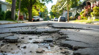 eine Straße, die sich durch eine Kleinstadt schlängelt. Die Straße weist deutliche Schäden auf, darunter mehrere Risse und Schlaglöcher.