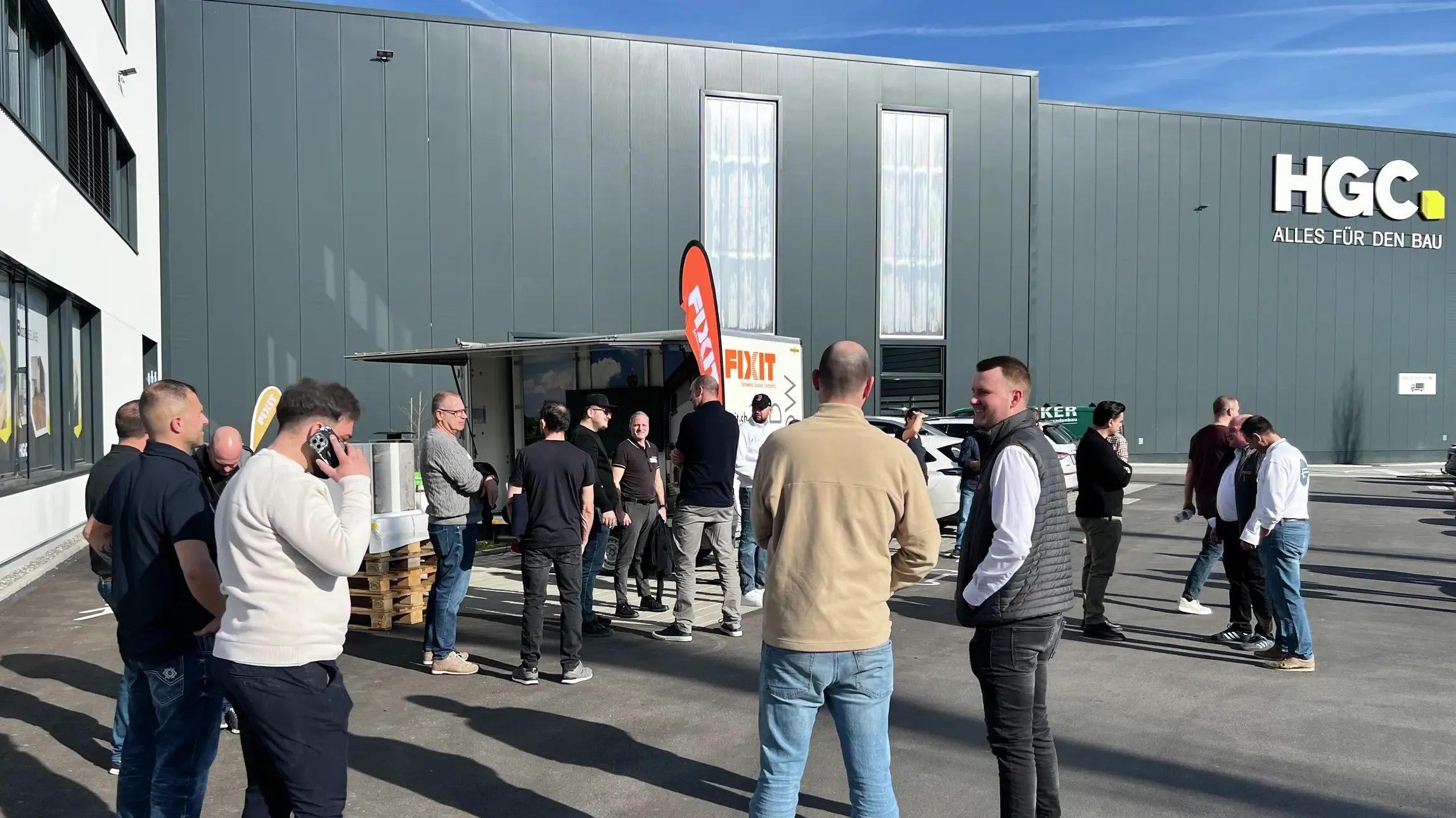 Aussenansicht der HGC Halle in Inwil mit vielen Teilnehmenden im Gespräch und dem Fixit Roadshow Anhänger und einer orangenen Beachflag