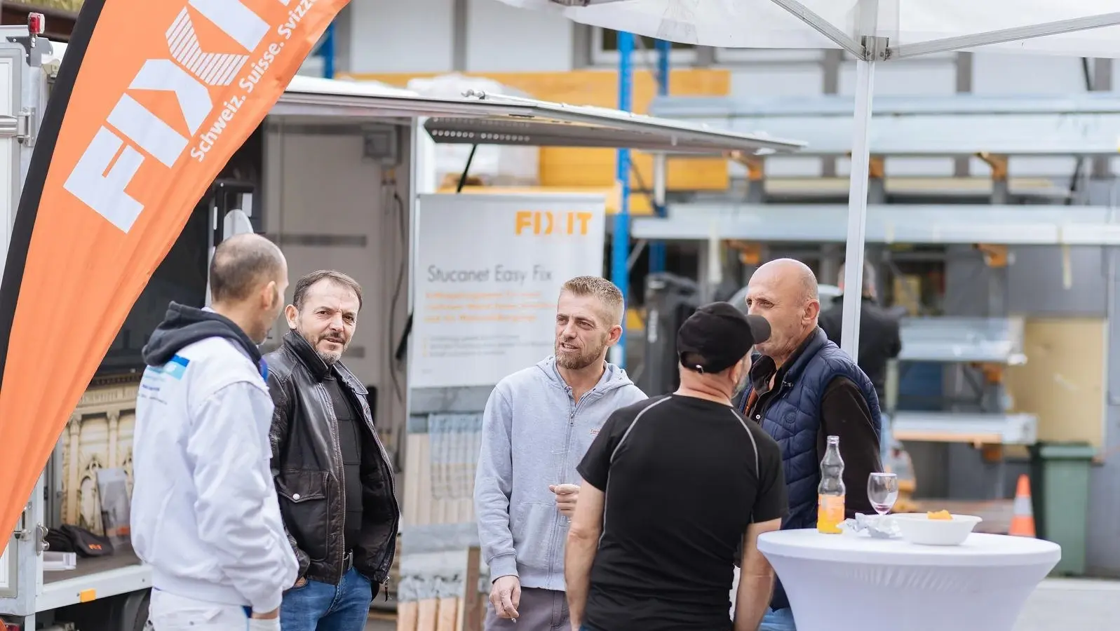 Drapeau orange Fixit Beach Flag lien, cinq personnes sont debout en cercle autour d'une table haute et discutent
