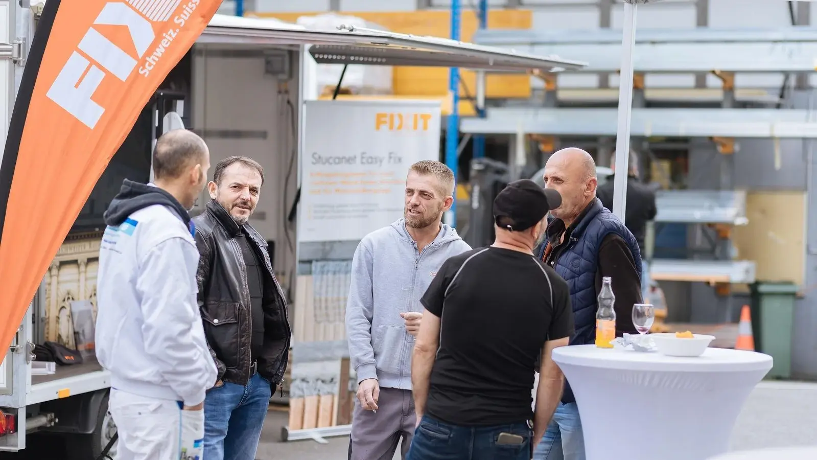 Drapeau orange Fixit Beach Flag lien, cinq personnes sont debout en cercle autour d'une table haute et discutent
