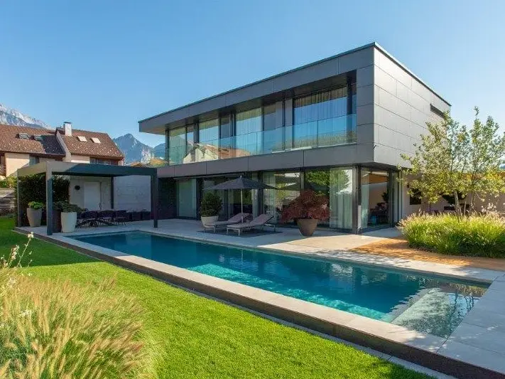 Un pré, une piscine rectangulaire et allongée devant une maison individuelle moderne de deux étages, baignée par la lumière du matin