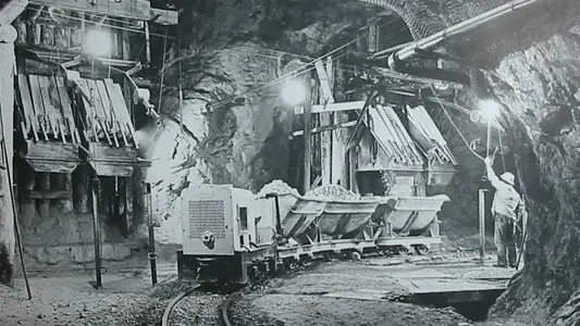 Photographie en noir et blanc d'une galerie avec des ouvriers, un train de wagonnets chargés de roches et l'installation de remplissage en arrière-plan